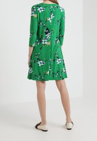 Zelené květinové šaty s tříčtvrtečními rukávy, mají rozšířenou sukni a složité květinové vzory v bílé, žluté a modré barvě.