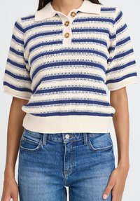 Prugasta pletena majica s kratkimi rokavi v kremni in mornarski modri barvi, z ovratnikom, tremi rjavimi gumbi in rebrastim robom, v kombinaciji z jeans hlačami.
