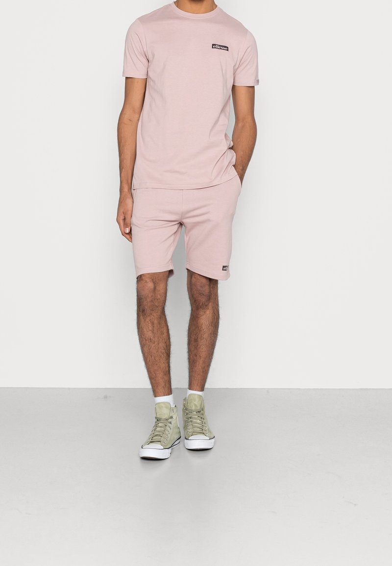 Homme portant une chemise à manches courtes rose clair et un short assorti avec des écussons de logo, des chaussettes blanches, et des baskets montantes beige, debout contre un mur blanc.