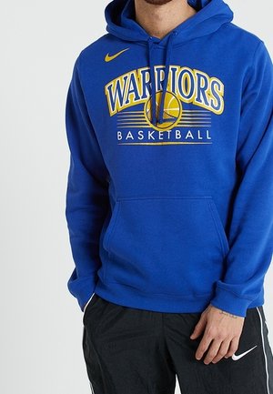 Sudadera azul con capucha con texto y logo de "Warriors Basketball", llevada con pantalones negros de Nike, mostrando parte de una persona de pie.