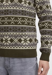 Strickpullover mit geometrischem Muster in Dunkelgrün, Grau und Creme, mit gerippten Bündchen und Saum, sowie einem Etikett auf der unteren Seite.