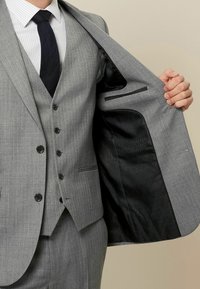 Sivi trodijelni odijelo s teksturiranim završetkom, koje uključuje sako s jednim kopčanjem, dugmadi i elegantnom crnom podstavom.