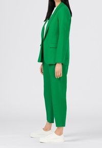 Zelený blejzr a odpovídající kalhoty vyrobené z hladkého materiálu. Blejzr má klopové límce; oblečení doplněné bílými pohodlnými teniskami.