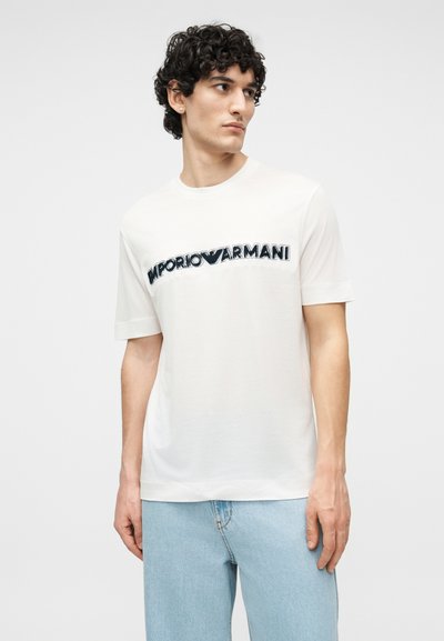 Νεαρός με κατσαρά μαλλιά που φοράει λευκό t-shirt Emporio Armani και ανοιχτόχρωμο τζιν, κοιτάζοντας προς τα πλάγια σε απλό φόντο.