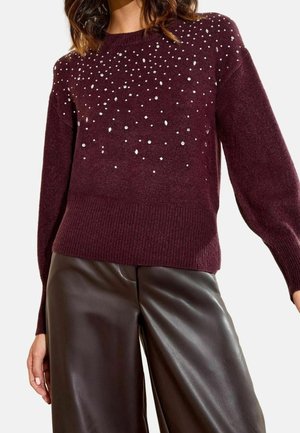 Kvinde iført en vinrød sweater med spredte små hvide pynt, kombineret med mørkebrune læderbukser med vide ben.