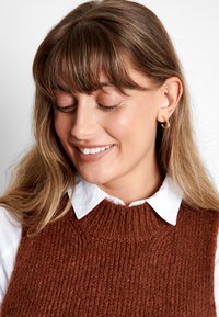 Bruine gebreide sweater vest met een gestructureerd patroon, gedragen over een witte blouse met kraag, met een hoge halslijn en geribbelde details.