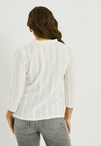 Blusa blanca con patrones texturizados intrincados y pequeñas aberturas, con un escote con volantes y mangas tres cuartos.