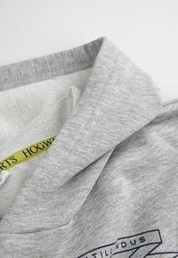 Szary bawełniany sweter z fałdowanym kołnierzem, wykonany z teksturowanej tkaniny, z żółtą metką wewnętrzną z napisem "HOGWARTS". Z ozdobnym nadrukiem graficznym.