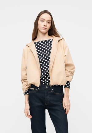 Ung kvinde med langt brunt hår iført en beige jakke med lynlås over en sort og hvid prikket top og mørkeblå jeans mod en ensfarvet baggrund.