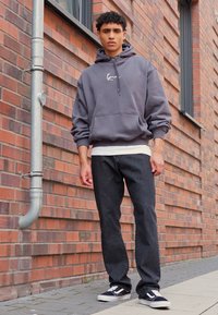 Grå hettegenser med frontlomme og snor, lagd over en hvit skjorte; kombinert med mørke jeans og svarte sneakers mot en mursteinsvegg.