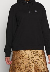 Czarna bluza z kapturem i regulowanym sznurkiem, z ribbowanymi mankietami i dolnym brzegiem, z małym białym logo "CK" na klatce piersiowej. Spódnica w złotym kolorze z czarnymi plamkami.