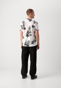 Korte mouwen wit T-shirt met zwart tropisch bloemenpatroon, gecombineerd met zwarte losse broek en zwarte schoenen, gezien van achteren.