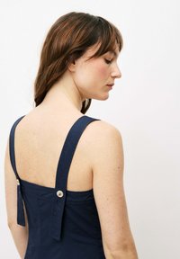 Das navyblaue Kleid verfügt über breite Träger, ein figurbetontes Oberteil und einen tiefen Rückenausschnitt mit zwei Knopfakzenten nahe der Schulter. Glattes, strukturiertes Material.