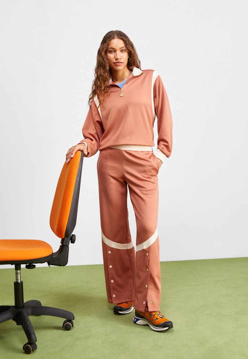Ensemble de sport en rose poussiéreux avec un col zippé, des accents crème et des boutons-pression sur le pantalon. Le modèle pose à côté d'une chaise de bureau orange.