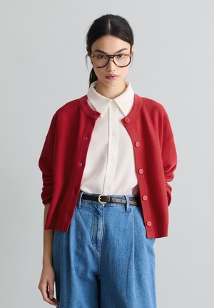 Giovane donna con capelli scuri e occhiali, indossa un cardigan rosso abbottonato sopra una camicia bianca con colletto e jeans blu a vita alta con cintura nera.