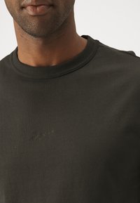 Černé bavlněné tričko s kulatým výstřihem a decentním detailem loga. Materiál je hladký, s přiléhavým střihem a minimem švů.