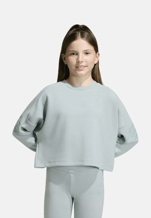 Joven chica con cabello largo lleva una sudadera azul claro de gran tamaño y leggings a juego, de pie frente a un fondo gris liso.