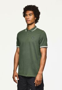 Uomo in piedi, che indossa una polo verde scuro con bordi bianchi su colletto e maniche, abbinata a pantaloni cargo neri, di fronte a uno sfondo chiaro e semplice.