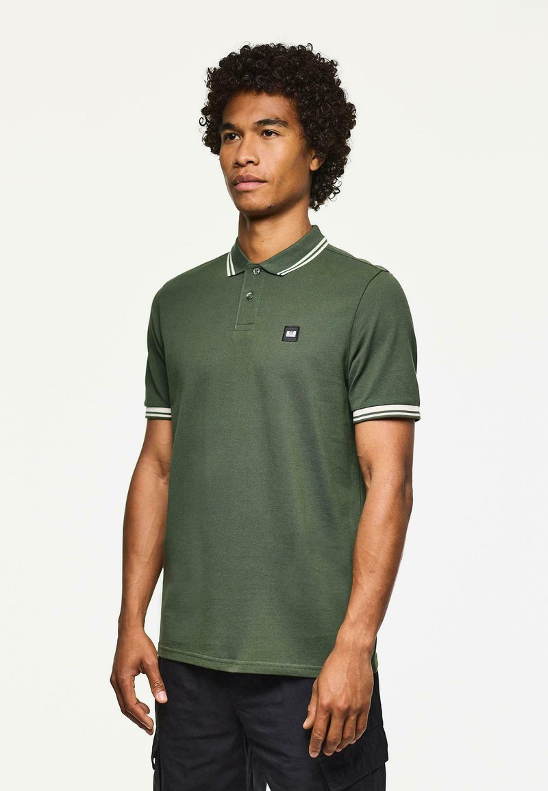 Uomo in piedi, che indossa una polo verde scuro con bordi bianchi su colletto e maniche, abbinata a pantaloni cargo neri, di fronte a uno sfondo chiaro e semplice.