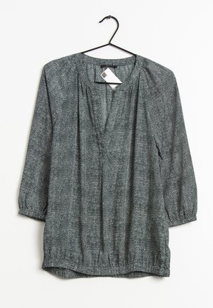 Blouse à manches longues tachetée noir et blanc avec poignets élastiques et fente à l'encolure, suspendue à un cintre noir sur fond blanc.
