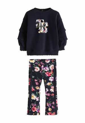 Junges Kind steht und trägt ein schwarzes, mit Blumen gemustertes Outfit mit langärmligem Oberteil und passenden Hosen sowie weißen Turnschuhen.