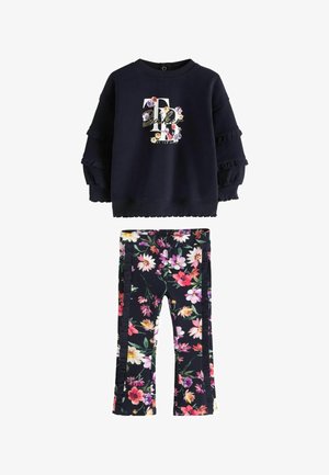 Lille barn står iført en sort blomsterprintet dragt med en langærmet top og matchende bukser samt hvide sneakers.