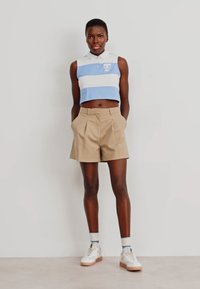Camiseta polo azul y blanca de rayas cortada, combinada con pantalones cortos khaki con pliegues y zapatillas blancas con suela beige, sobre un fondo neutral.