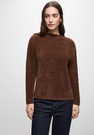 Pitkähiuksinen nainen, jolla on päällään ruskea pörröinen pitkähiha-paita ja tummansiniset farkut, seisoo yksinkertaisen vaalean taustan edessä.