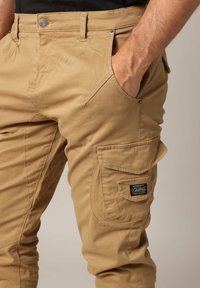 Beige cargo broek gemaakt van stevig materiaal, voorzien van een knoopsluiting, zijzakken en een opvallend label op de onderste zak.