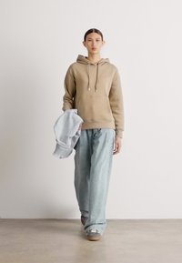Bež dukserica s vezicama i prednjim džepom, uparena s laganim plavim oversized trapericama. Model drži svijetlu traper jaknu, nosi sive tenisice.