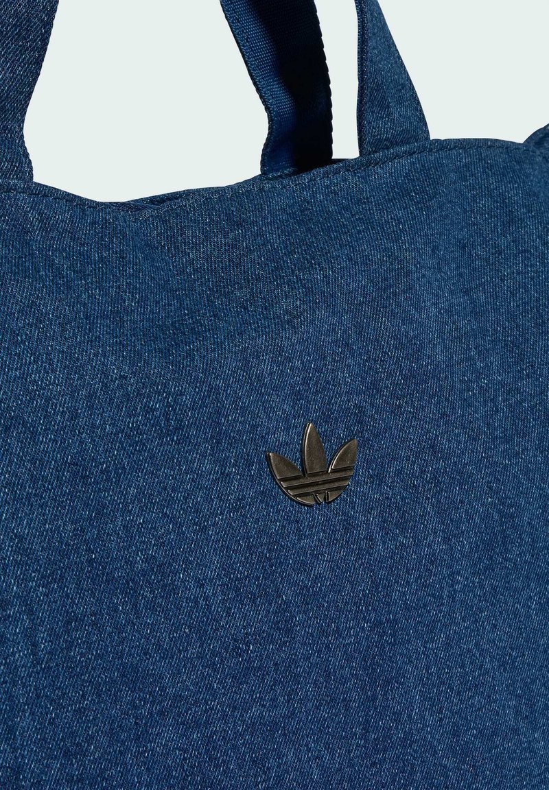 Denim tote bag in dark blue with a textured surface. Features two fabric handles and a black metal logo accent on the front.