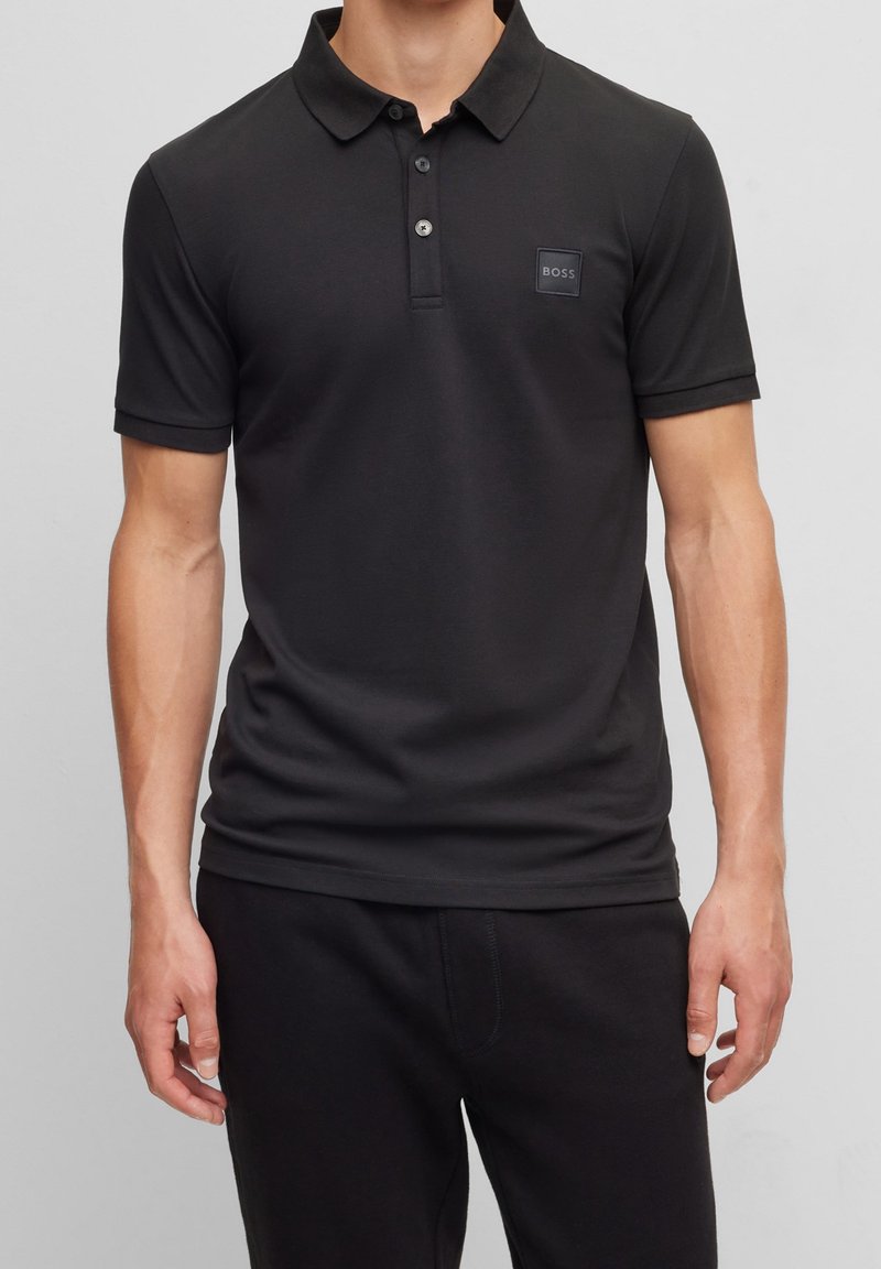 Homme portant un polo noir à manches courtes avec un col et trois boutons, avec un petit logo BOSS sur la poitrine.