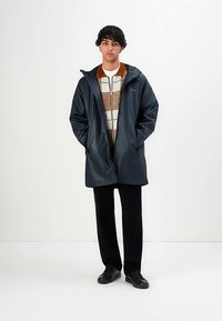 Laivastonsininen vedenpitävä takki hupulla, päällä kuviollinen neulepusero. Malli seisoo valkoista taustaa vasten, kädet taskuissa.