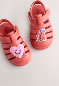 Rote Gummisandalen mit ausgeschnittenem Design, verziert mit einem lächelnden Herz und Marienkäfer-Applikationen auf den Riemen. Weiche Textur, verstellbarer Verschluss.