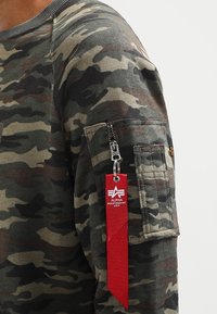Camicia a maniche lunghe con motivo mimetico in tonalità verdi e marroni, dotata di tasca con zip sul braccio e di un'etichetta rossa che mostra il logo del marchio.