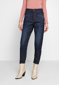 Jeans de mezclilla azul oscuro con cintura alta, pierna recta y sutiles efectos de desgastado. Combinados con botas de tacón beige.