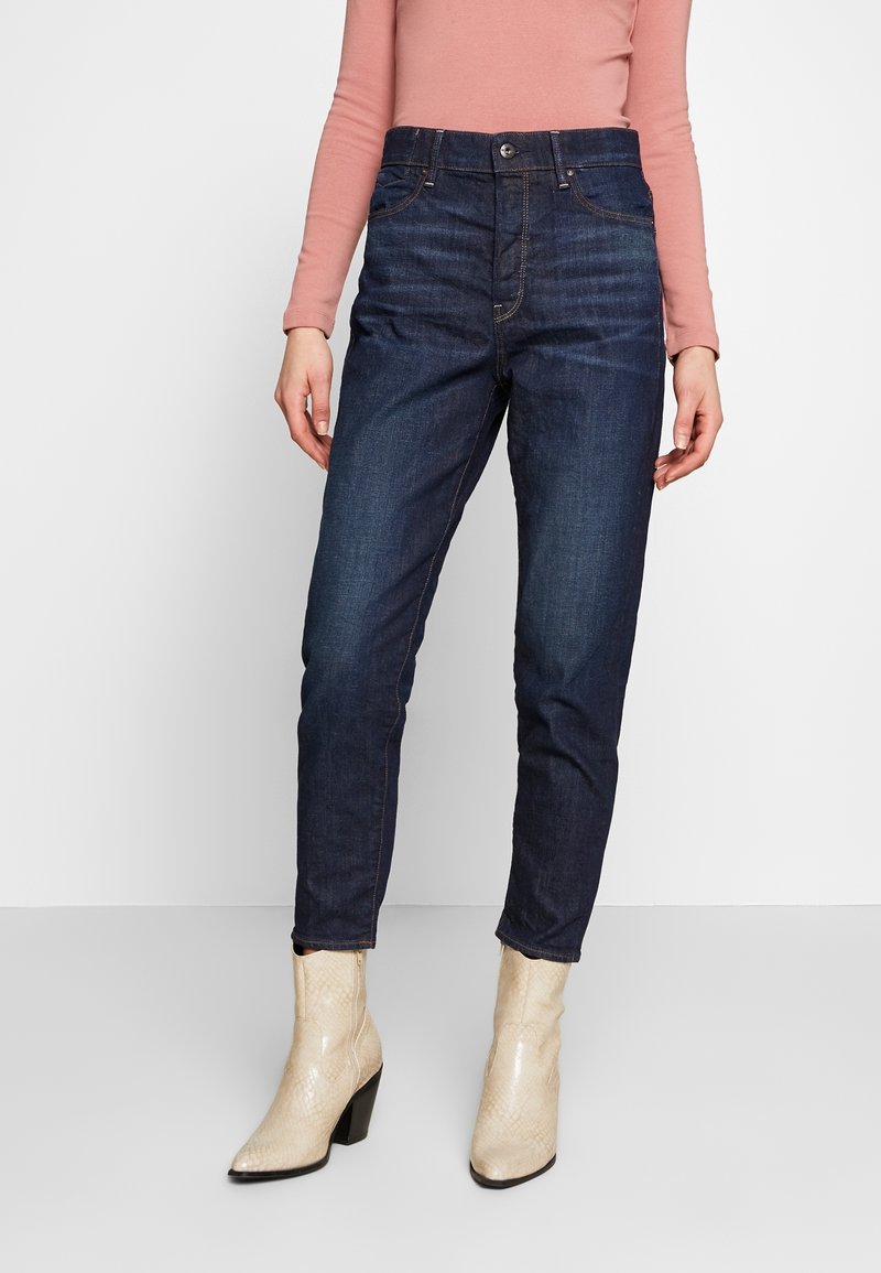 Jeans de mezclilla azul oscuro con cintura alta, pierna recta y sutiles efectos de desgastado. Combinados con botas de tacón beige.