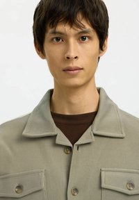 Jonge man met kort, donker haar, die een beige knoopshirt over een kastanjebruine ronde halstrui draagt, recht vooruit kijkend met een neutrale expressie.