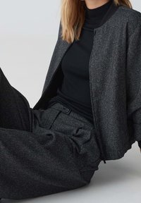 Femme assise portant une veste bomber gris foncé texturée et un pantalon large assorti avec un haut à col roulé noir.