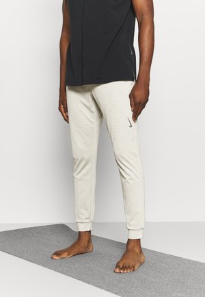 Jasnoszare spodnie sportowe o teksturowanym wykończeniu i elastycznych mankietach, połączone z czarnym bezrękawnikiem, noszone boso na szarej macie.