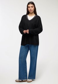 Kobieta w czarnej dzianinowej bluzie na białej bluzce z koronkowym wykończeniem, w niebieskich szerokich dżinsach i białych trampkach z gumową podeszwą, na białym tle.