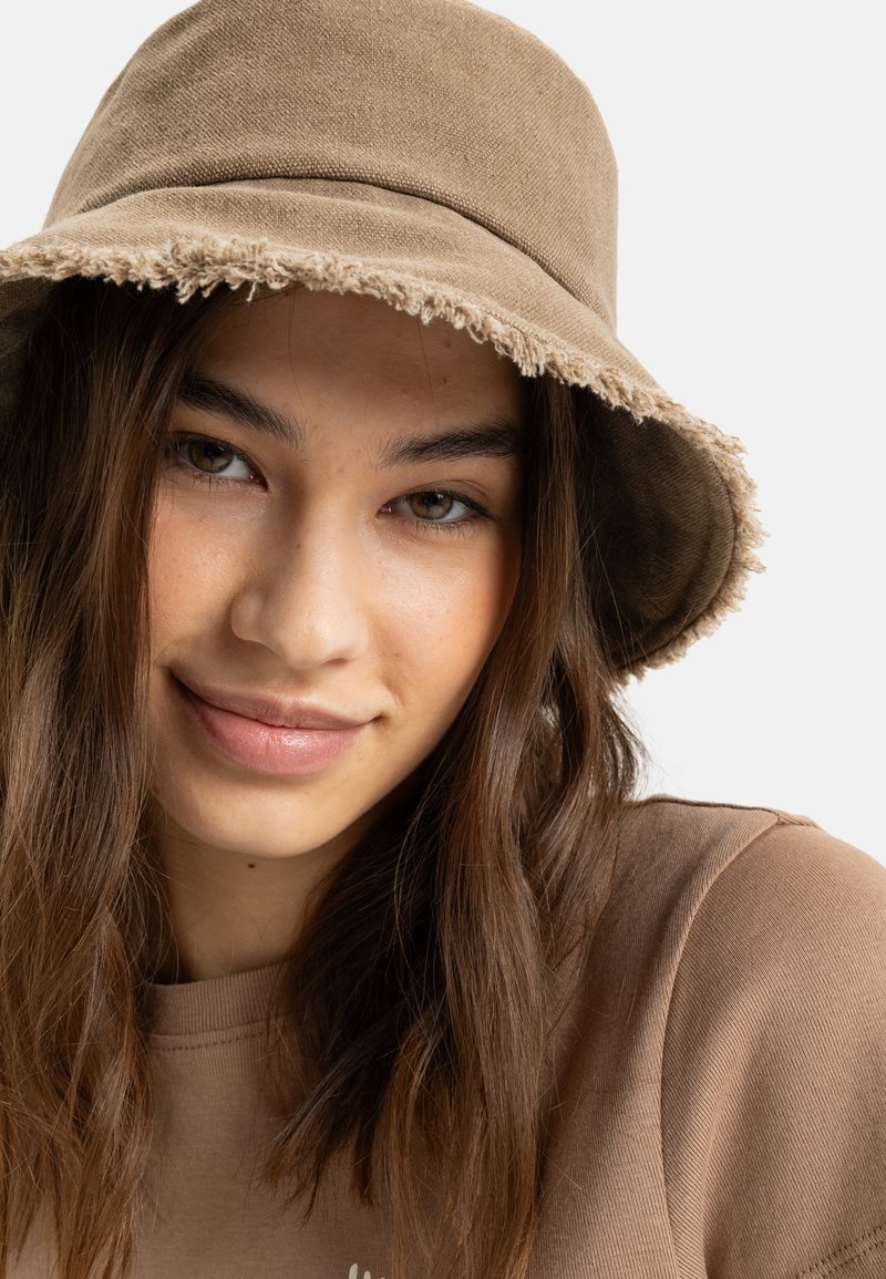Brun bomuldsbucket hat med rå kant, parret med en matchende brun top. Glat tekstur, enkelt design og afslappet udseende.