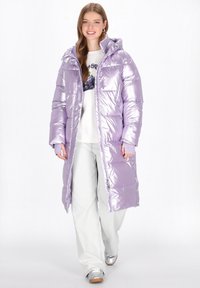 Hosszú, fényes levendula színű puffermellény kapucnival, steppelt szegmensekkel, két elülső zsebbel és teljes hosszúságú cipzárral. Fehér felsővel párosítva.