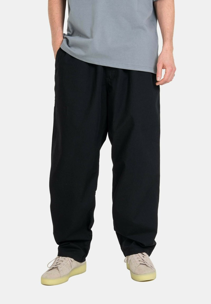 Schwarze Baumwollhosen mit lockerer Passform, ausgestattet mit einem elastischen Taillenbund und seitlichen Taschen, kombiniert mit hellbeigen Sneakers.