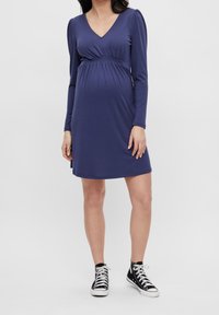 Kvinna klädd i en långärmad marinblå gravidklänning med empiremidja och svarta högskaftade sneakers mot en enkel bakgrund.