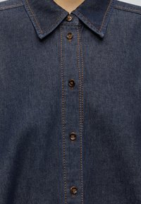 Chemise en denim bleu foncé avec des boutons marron et des coutures orange, montrant le col et la patte de boutonnage à l'avant.
