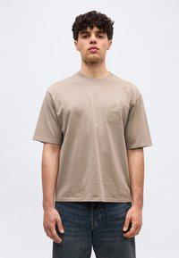 Νεαρός άνδρας με σγουρά μαλλιά, φοράει μπλουζάκι σε μπεζ χρώμα, oversized, με διακριτική κεντητική στο στήθος και σκούρο μπλε τζιν, στέκεται μπροστά σε απλό φόντο.