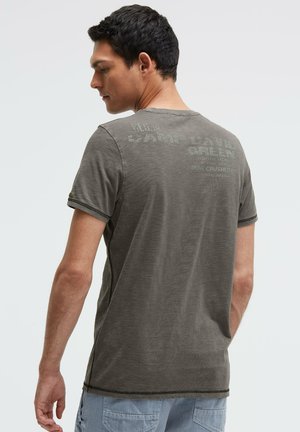 Man facing away wearing a faded dark gray graphic T-shirt and light blue shorts against a plain light background.