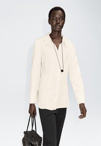 Lysebeige skjorte med knapper, lange ærmer, spidst krave og sideslidser, kombineret med sorte bukser og en sort taske med struktur.