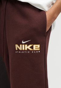 Άτομο που φοράει καφέ αθλητικές φόρμες Nike με το λογότυπο «NIKE ATHLETIC CLUB» σε κίτρινο και λευκό, με το χέρι να ακουμπάει μέσα στην τσέπη.
